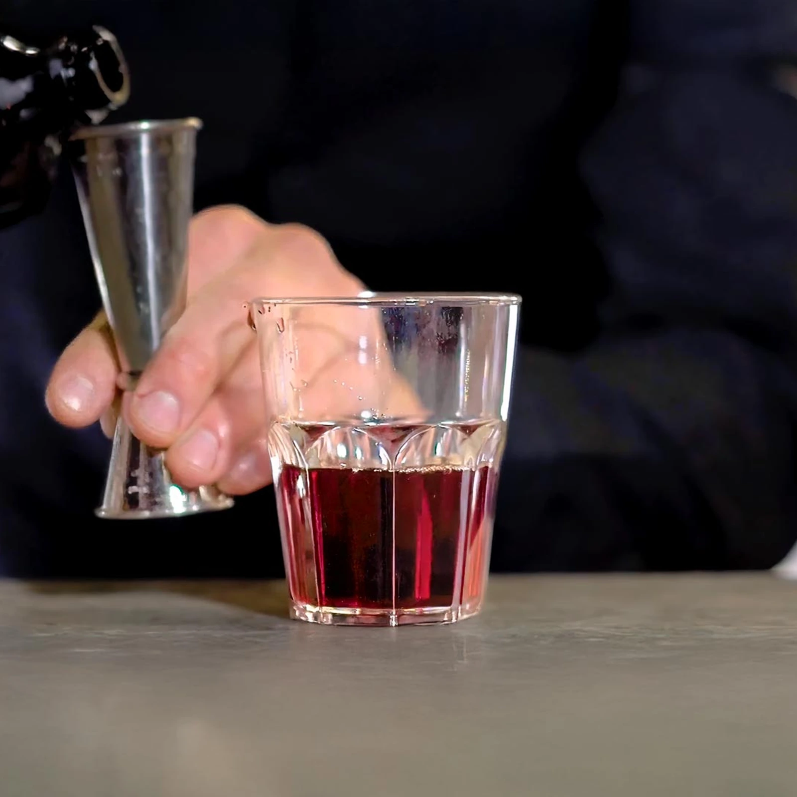 Mehrwegbecher Granity Clear 290 ml auf dem Tresen, der Barkeeper misst einen roten Drink mit dem Jigger ab