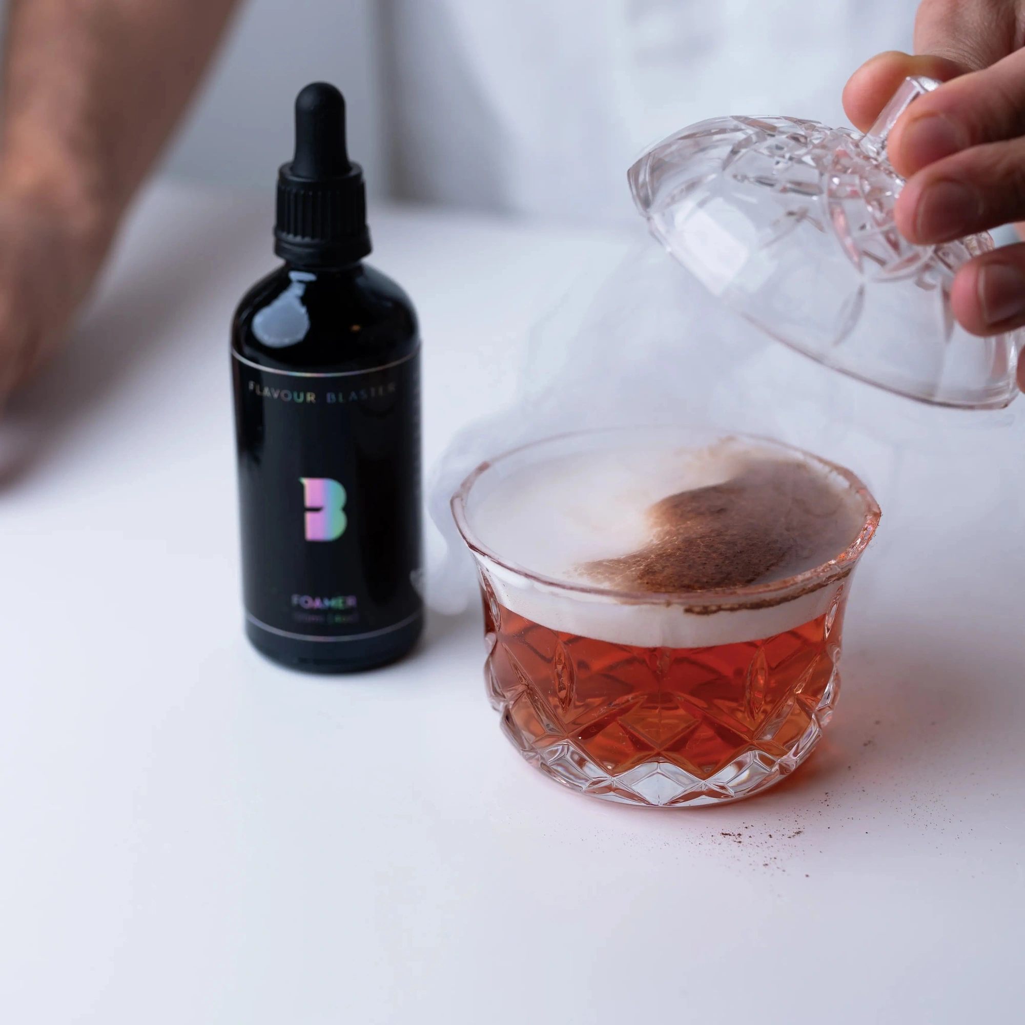 Foamer cocktail Flavour Blaster: αφρός πάνω σε κόκκινο κοκτέιλ σε κρυστάλλινο ποτήρι με σκαλιστό σχέδιο, η γυάλινη καμπάνα σηκωμένη και το μπουκάλι δίπλα